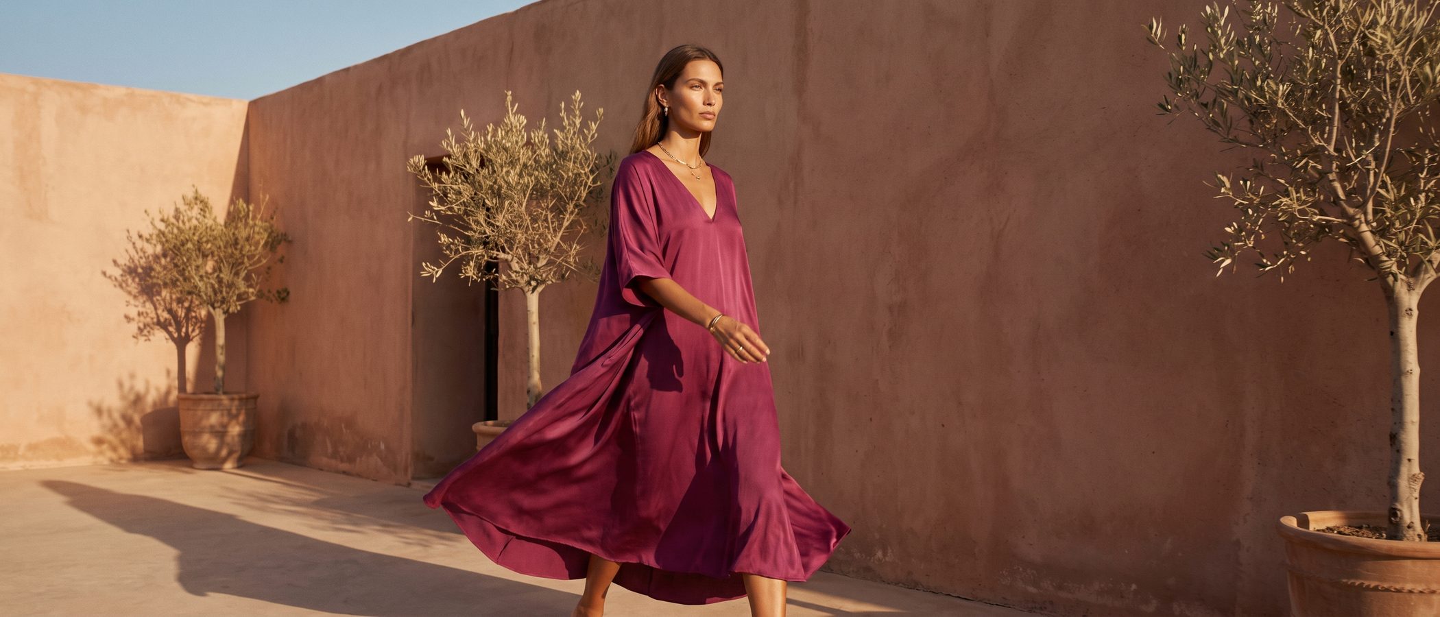 Campagnebeeld: model in een wijde magenta zijden jurk loopt in laag gouden zonlicht langs een terracotta muur met olijfboompjes
