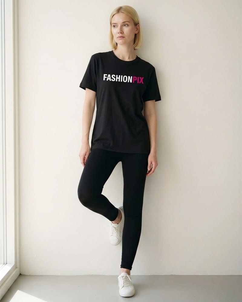 Iris, een jonge blonde vrouw, leunt bij raamlicht tegen een witte muur in het FASHIONPIX-T-shirt en kijkt van de camera weg
